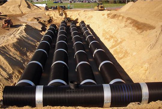 Installation of large black drainage pipes underground at a construction site, surrounded by sand and machinery.
