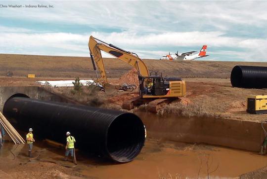 Construction site with workers installing large black pipes, an excavator, and an aircraft in the background.