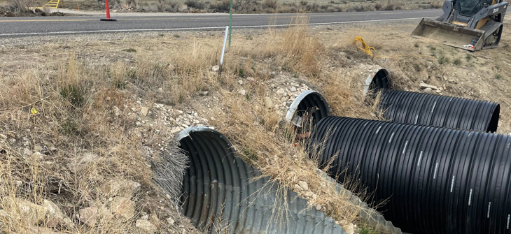 Corrugated metal and plastic culvert pipes are installed in a roadside drainage ditch near a rural highway.