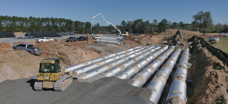 Construction site with large metal culvert pipes being installed in parallel, with machinery and workers present.