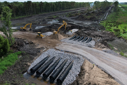 Construction site with excavators installing large drainage pipes and a partially cleared forest area in the background.