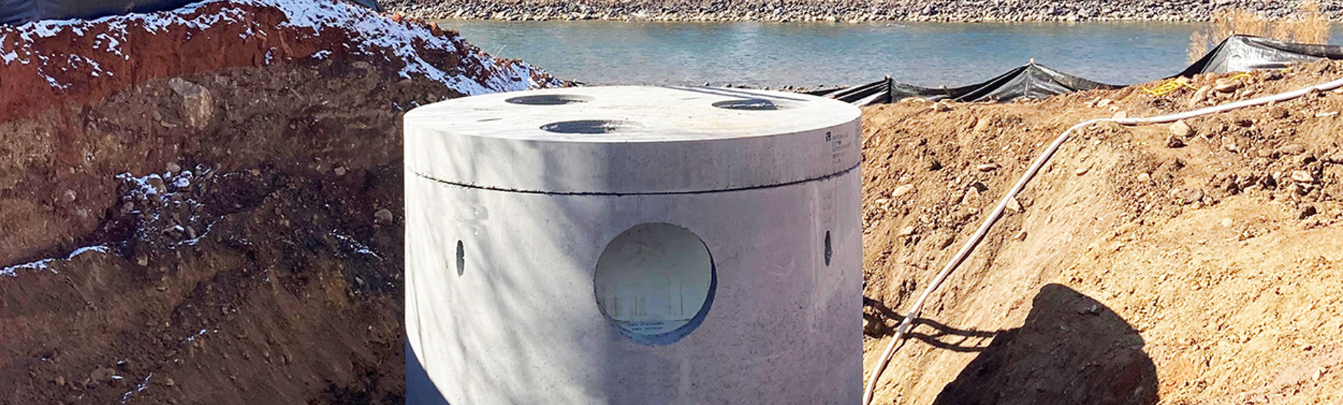 Concrete manhole structure installed in a dug trench near a river with hills and buildings in the background.