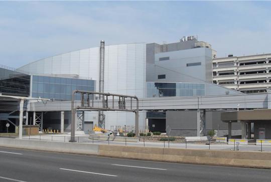 Modern industrial building with glass and metal facade, adjacent to a multi-level parking structure, next to a roadway.