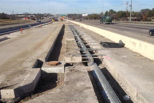 Construction site on a highway with concrete barriers and exposed rebar. Cars are visible on the adjacent operational lanes.