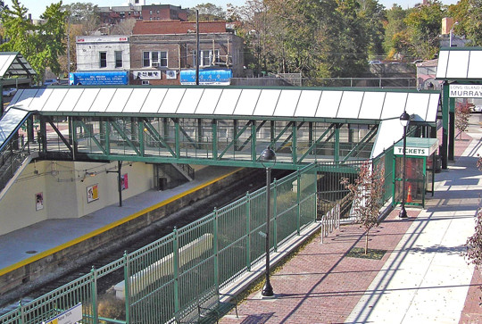 Murray Hill train station with a green pedestrian overpass, platform, ticket area, and surrounding trees and buildings.