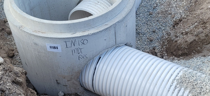 Large concrete drainage structure with white corrugated pipes set in gravel at a construction site.
