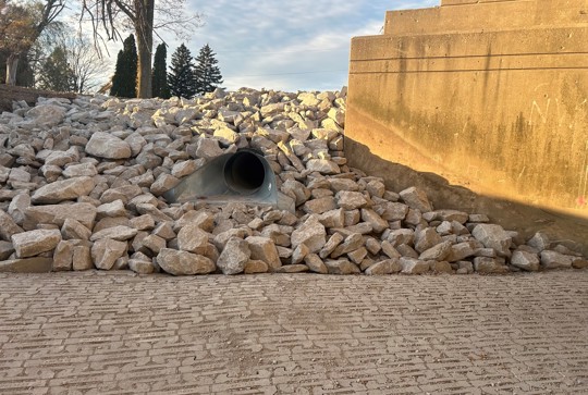 A drainage pipe is embedded in a rocky embankment, next to a concrete structure, under a partly cloudy sky.
