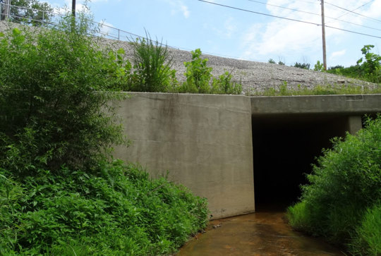 Concrete drainage tunnel entrance surrounded by greenery, under a gravel slope with overhead utility lines.