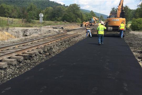 Construction workers pave a path alongside railroad tracks using heavy machinery, with a scenic backdrop of trees and hills.