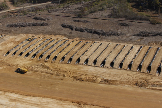 Aerial view of a landfill with rows of waste covered by soil, surrounded by cleared land and construction vehicles.