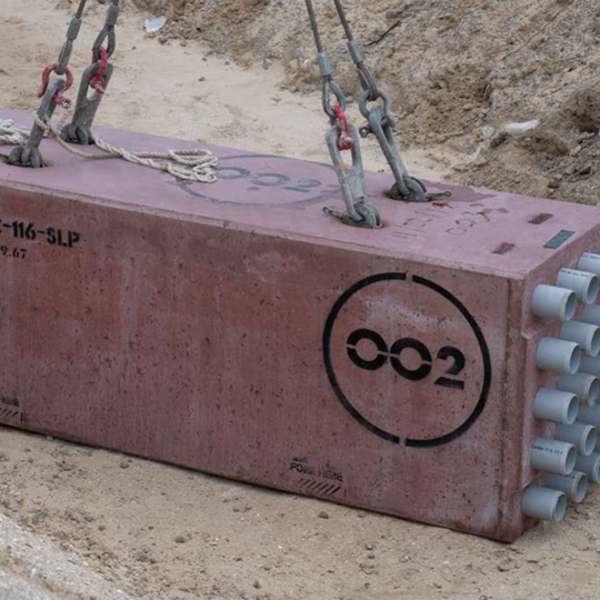 Large rectangular concrete block with multiple PVC pipe conduits being lifted by cables at a construction site.