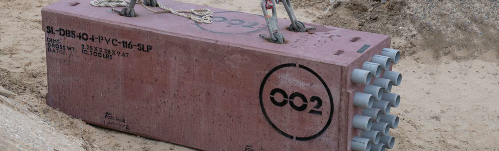 Large rectangular concrete block with multiple PVC pipe conduits being lifted by cables at a construction site.
