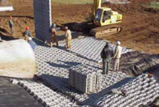 Construction site with workers placing interlocking concrete blocks and a yellow excavator in the background.