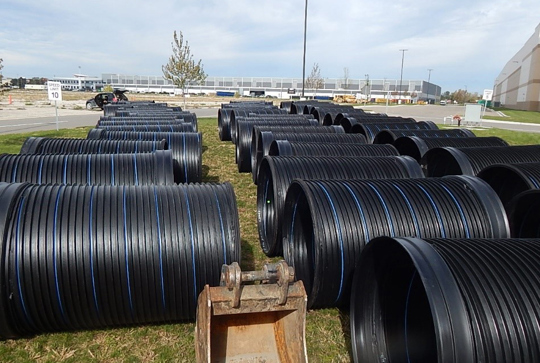 Rows of large black drainage pipes are stacked on grassy ground, with a construction site and industrial building in the background.