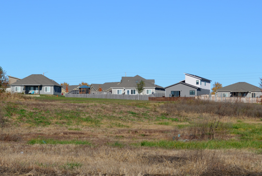 Suburban neighborhood with houses and fenced yards, surrounded by open grassy fields under a clear blue sky.