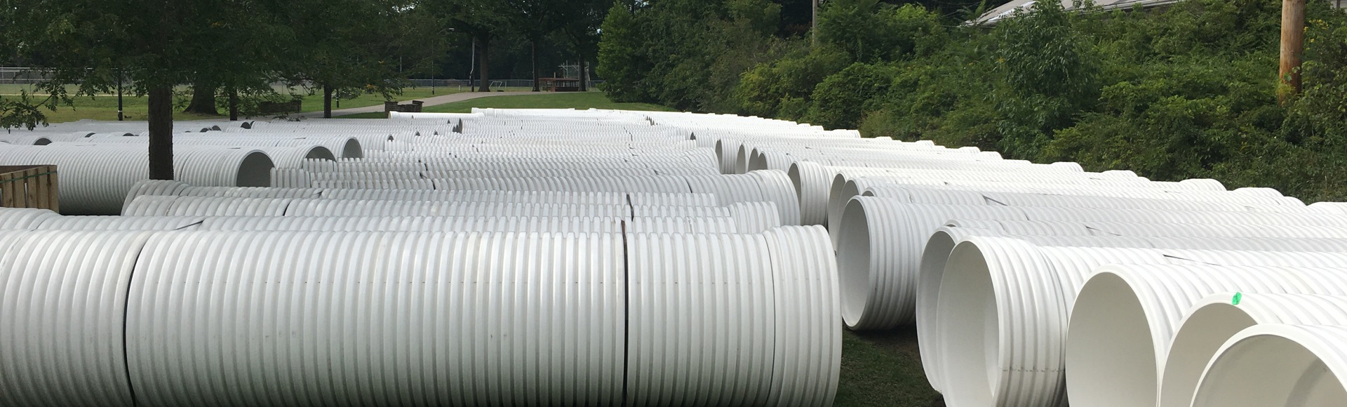 Large white corrugated plastic pipes are stacked in rows on grass near trees and power lines outdoors.