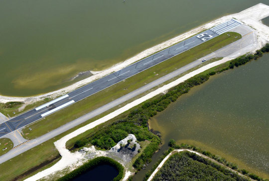 Aerial view of a runway extending into a body of water, surrounded by greenery and a small road on the left.