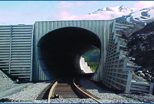 Train tracks leading into a metal tunnel entrance, surrounded by rocky terrain with a mountain in the background.