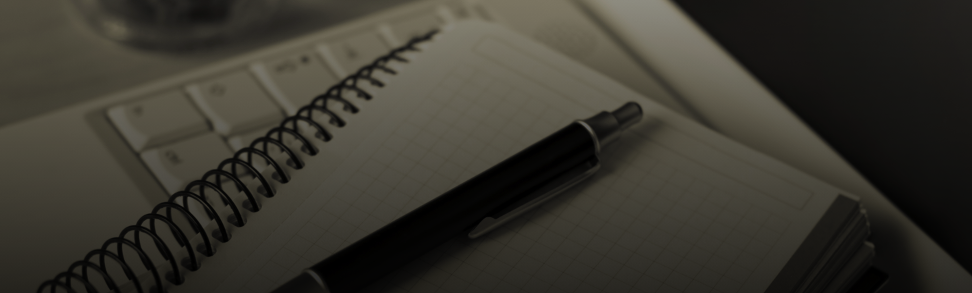 A spiral notebook and pen rest on a laptop keyboard next to a tablet.
