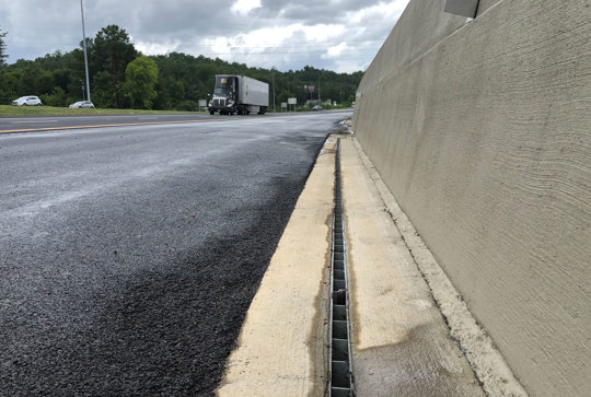 Close-up view of a roadside drainage system beside a paved road with a truck approaching in the background under cloudy skies.