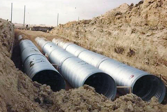 Three large metal pipes partially buried in a trench surrounded by dirt and earthworks under cloudy skies.
