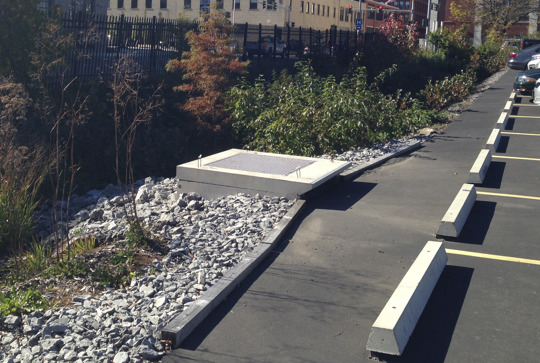 A parking lot with white barriers on the right and a sidewalk with piled rocks and vegetation on the left. A concrete slab is visible.