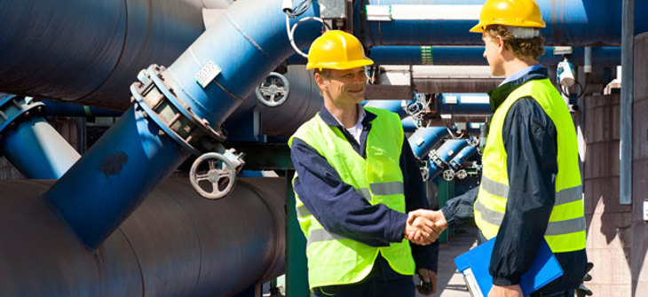 Two workers in safety vests and helmets shake hands in an industrial plant with large pipes in the background.