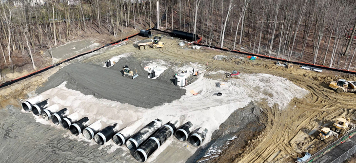 A construction site with large concrete pipes, earth-moving equipment, and cleared dirt near a wooded area.