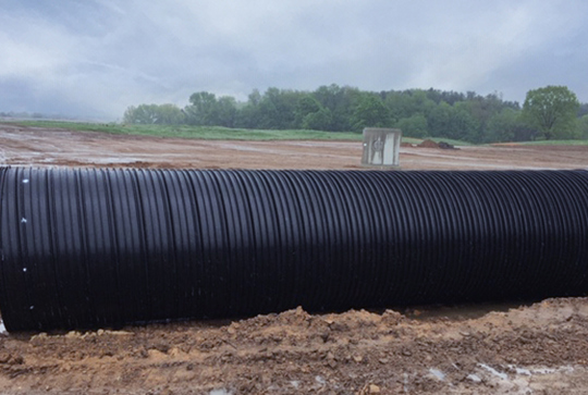 Large black corrugated pipe lies on muddy ground at a construction site with cloudy sky and trees in the background.