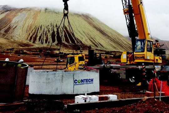 Construction site with a crane lifting a concrete structure. Workers are present, and a mountain is visible in the background.