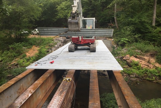 An excavator places metal sheets on a bridge framework over a stream in a forested area.