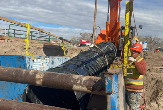 Construction worker installing a large black pipe with machinery at a construction site on a clear day.