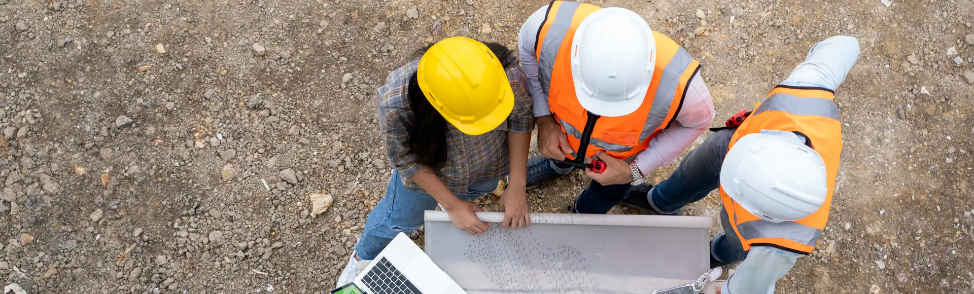 Three people in safety gear review plans on a table next to a laptop at a construction site, viewed from above.