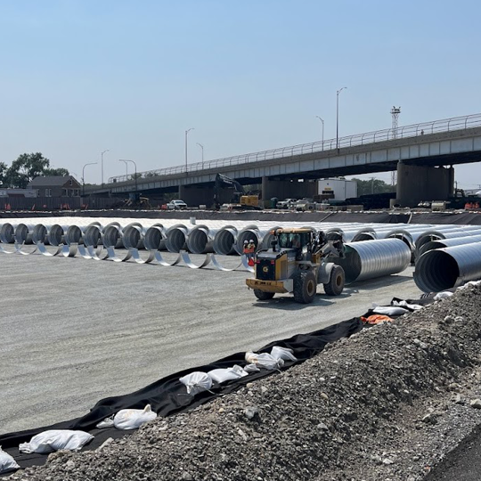 Construction site with rows of large metal pipes on a gravel area, forklift moving pipes, bridge and vehicles in background.