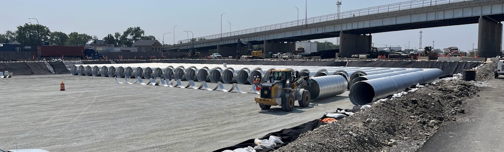 Construction site with rows of large metal pipes on a gravel area, forklift moving pipes, bridge and vehicles in background.