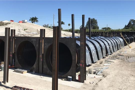 Large metal pipes and panels are arranged in a construction area, flanked by vertical steel beams, under a clear sky.