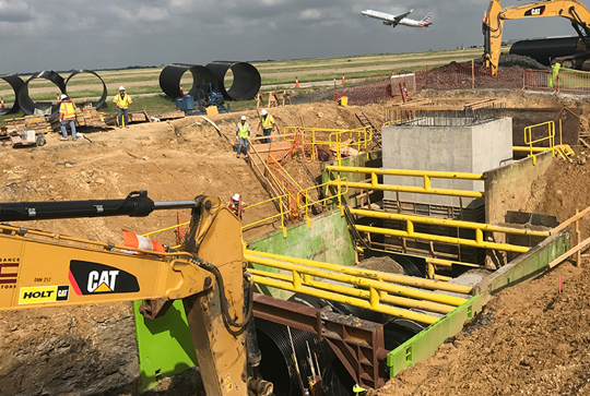 Construction site with heavy machinery and workers. Large pipes are visible, and a plane is taking off in the background.