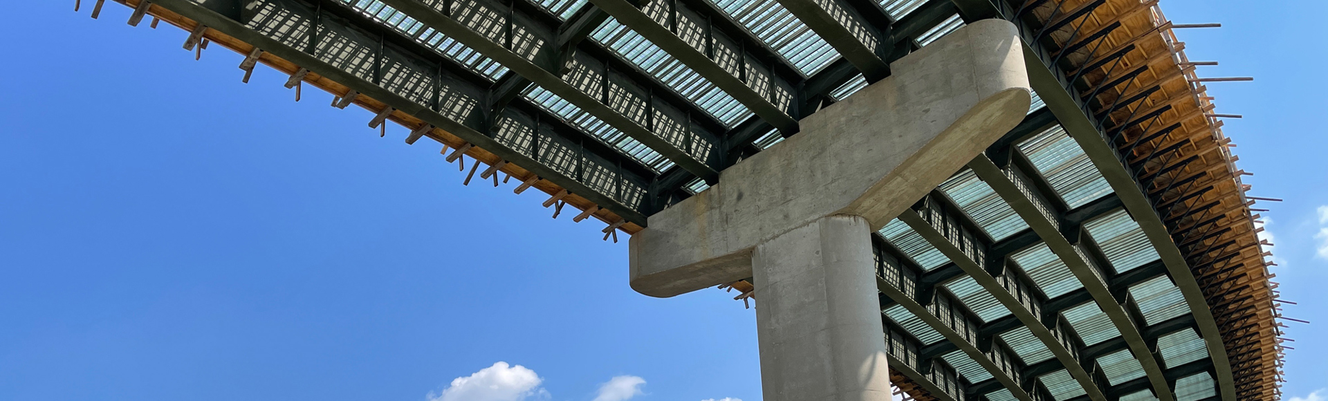 View from below of a modern bridge under construction, showing steel beams, concrete supports, and a clear blue sky.