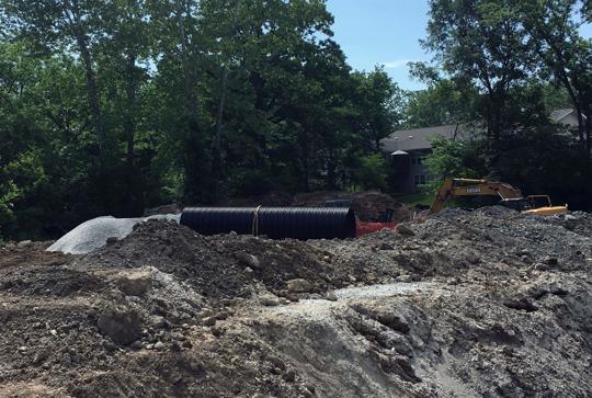 Construction site with excavated dirt, large pipes, and an excavator. Trees and a building are visible in the background.