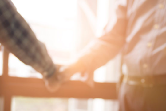 Two people shaking hands; one holding a yellow construction helmet. A computer is visible in the foreground.