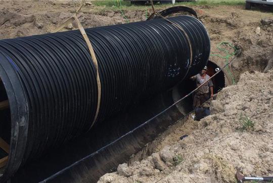 A large black pipe is being installed in a trench, with workers nearby and tools scattered around the area.