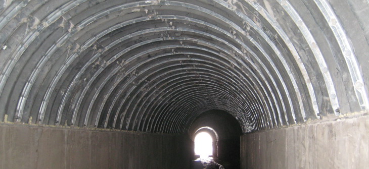 Inside view of a concrete and metal tunnel with light visible at the far end and a small stream running through it.