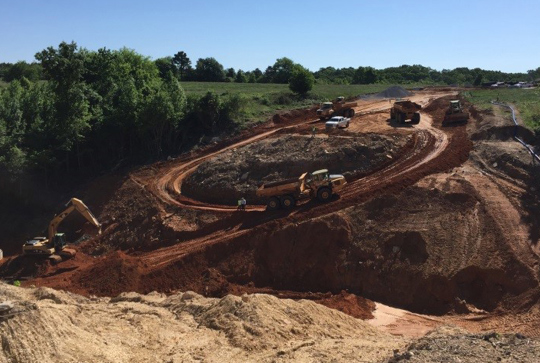 A construction site with trucks and excavators working on a dirt hill under a clear blue sky.