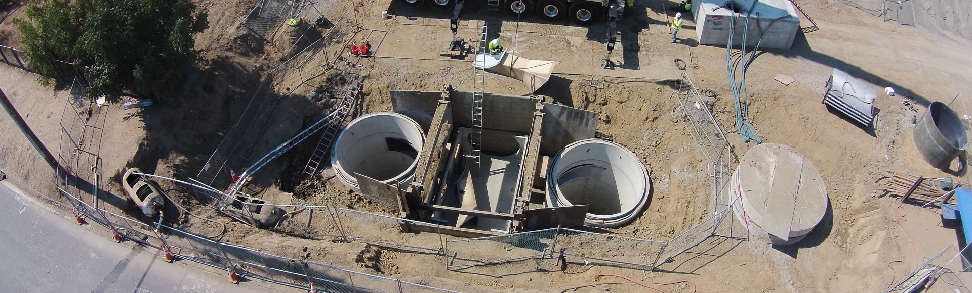 Overhead view of a construction site with two large circular concrete structures, a crane, and surrounding fencing.