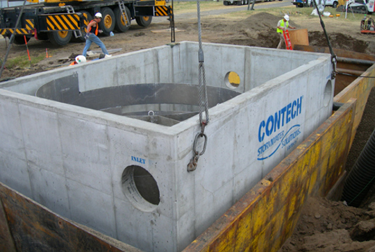 Concrete stormwater management structure being lowered into a construction pit by a crane, with workers nearby.